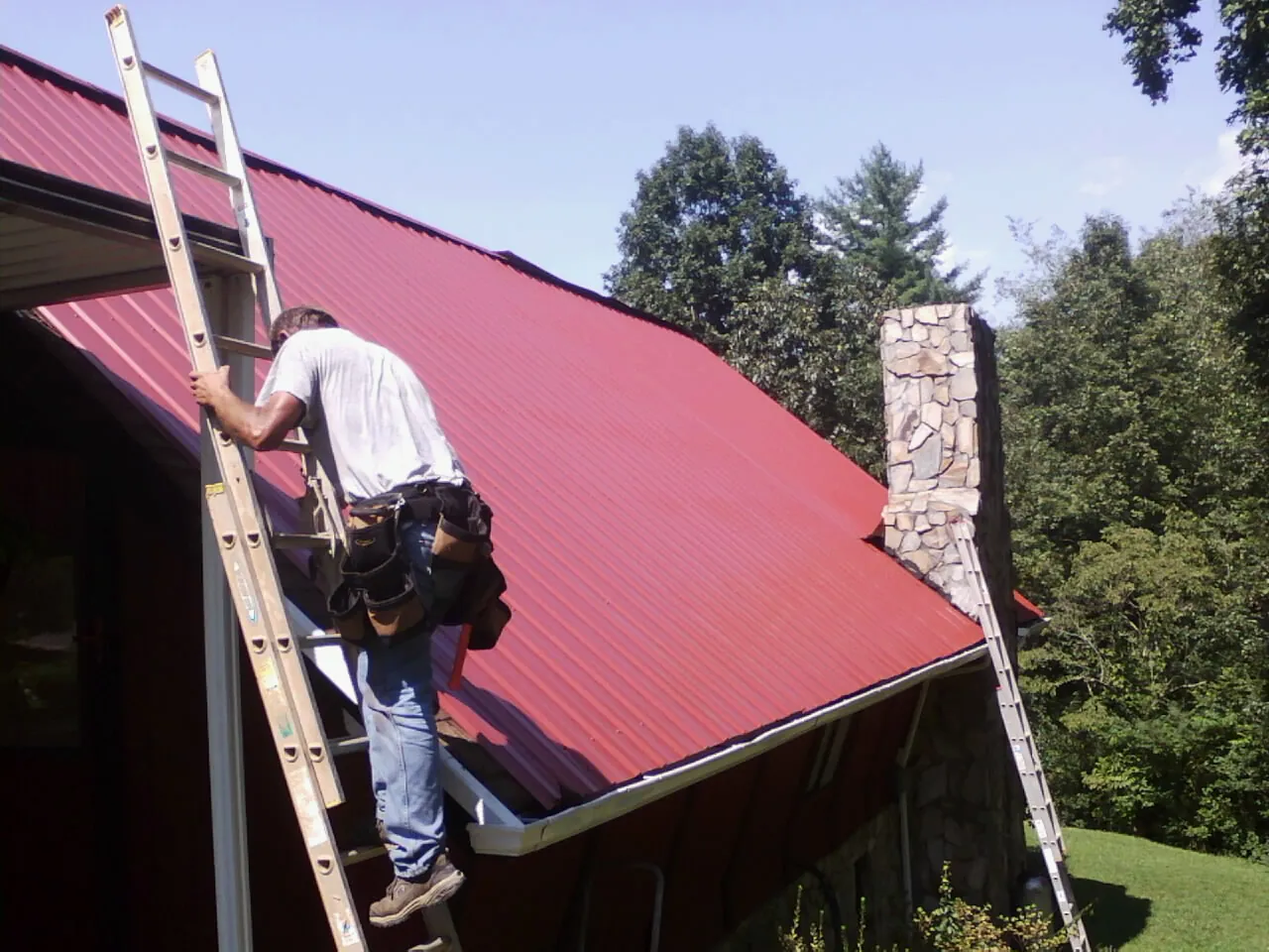 Roofer on a ladder beside a red metal roof and stone chimney — Soffit & Fascia Repair by Roofing Brothers in Kings Bay